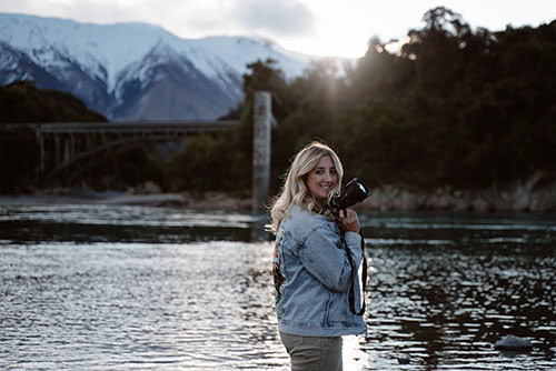 Photo of Kara in denim jacket by a river holding a camera.